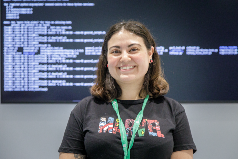 Picture of Tamara grinning, standing in front of a screen showing some coding. Tamara is a Software Developer for EMBL-EBI