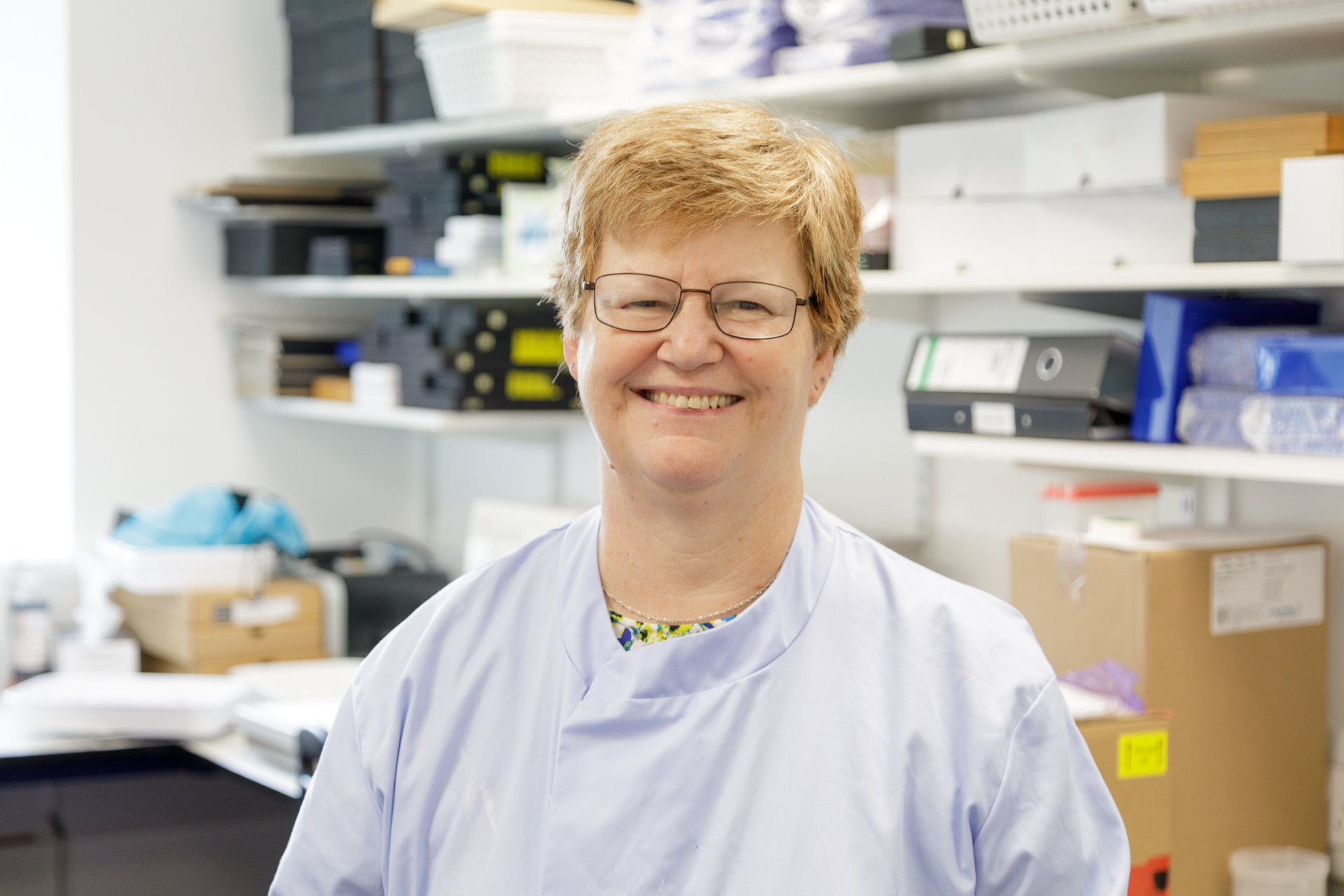 Picture of Yvette smiling. Yvette is in her lilac lab coat standing in the corner of a busy office/lab space.