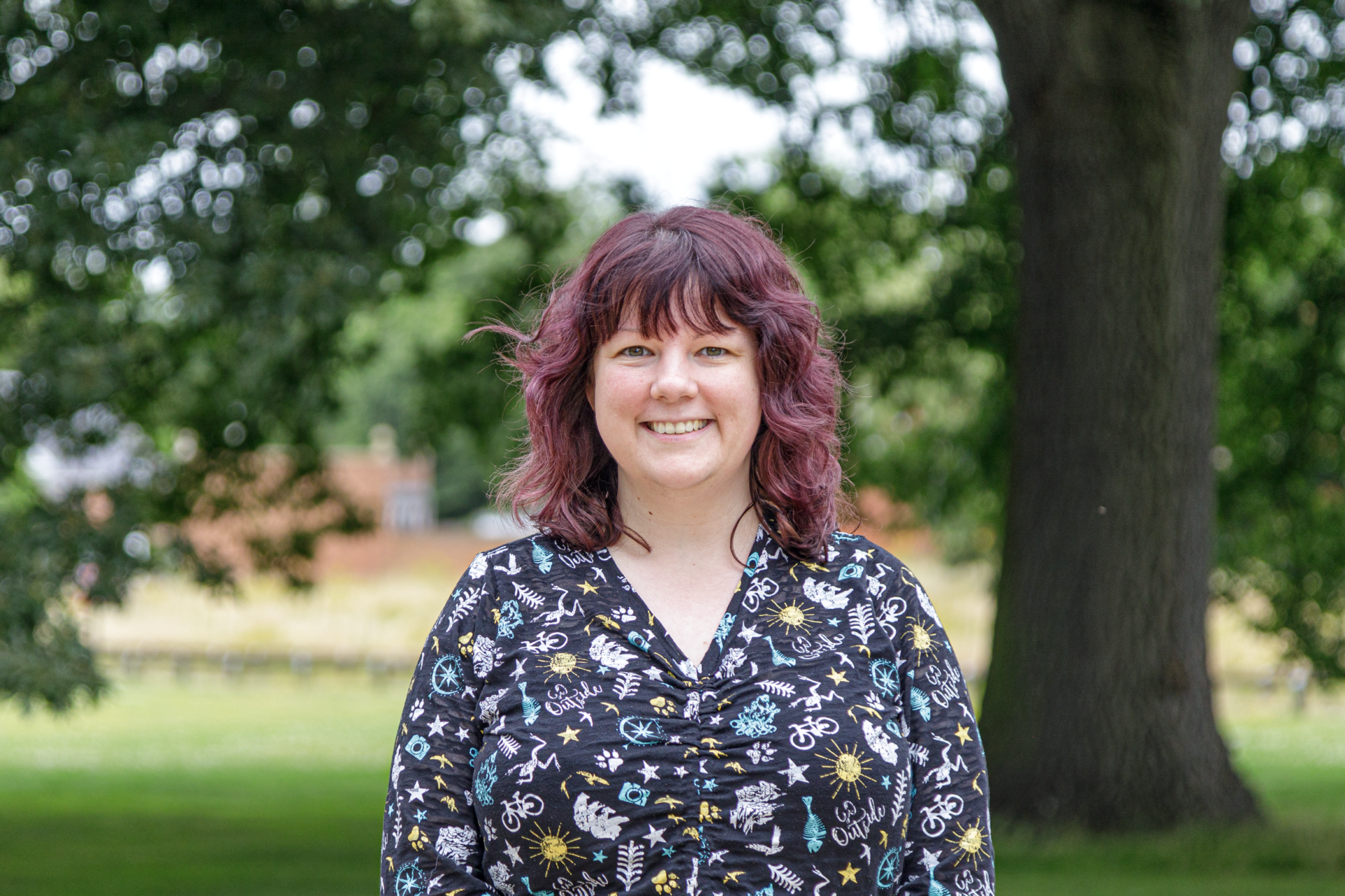 A picture of Jess smiling, standing in front of a large tree, Hinxton Hall is in the far background. Jess is a Research Governance Assistant at Wellcome Sanger Institute.